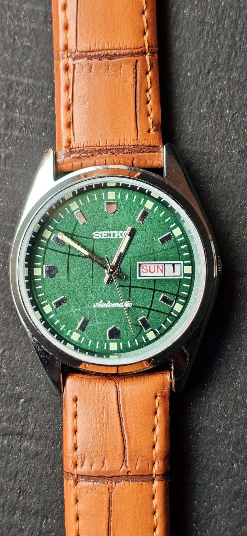 May include: A vintage Seiko automatic wristwatch with a green dial and a brown leather strap. The watch face displays the day and date, with the day reading "SUN 1". The watch has a silver-colored case.