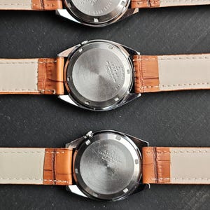 May include: Four vintage Seiko watches with brown leather straps are displayed. The watch backs are visible, showing stainless steel and water-resistant features. The straps are a light tan color.