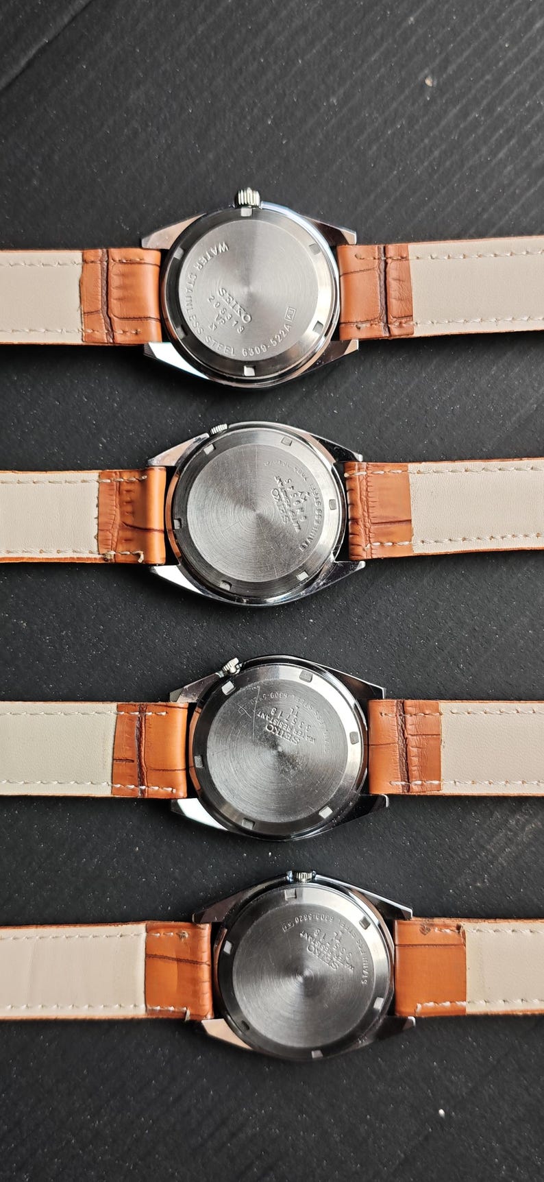 May include: Four vintage Seiko watches with brown leather straps are displayed. The watch backs are visible, showing stainless steel and water-resistant features. The straps are a light tan color.