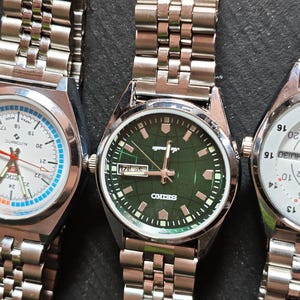 May include: Three stainless steel wristwatches with silver link bands. The watch faces are white, green, and white, with various markings and hands. The watches are displayed on a dark surface.