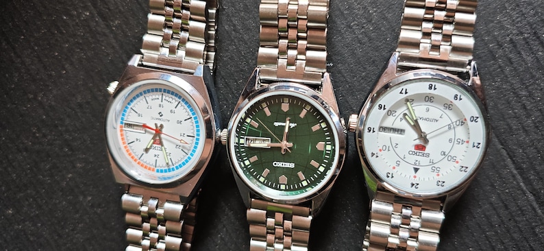 May include: Three stainless steel wristwatches with silver link bands. The watch faces are white, green, and white, with various markings and hands. The watches are displayed on a dark surface.