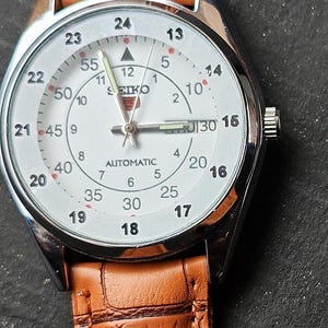 May include: A silver-toned Seiko automatic wristwatch with a white face and brown leather band. The watch face displays the time with black numerals and a second hand. The band has a textured, crocodile-like pattern.