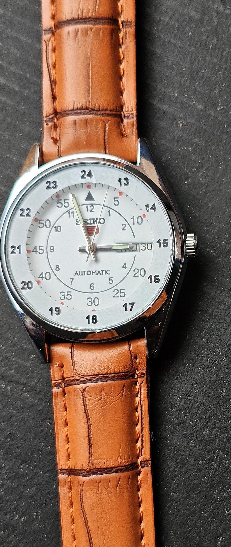 May include: A silver-toned Seiko automatic wristwatch with a white face and brown leather band. The watch face displays the time with black numerals and a second hand. The band has a textured, crocodile-like pattern.