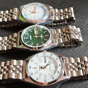 May include: Three stainless steel wristwatches with silver link bands are displayed. The watch faces include a white face with black numbers, a green face with silver hands, and a white face with black numbers and a red logo.