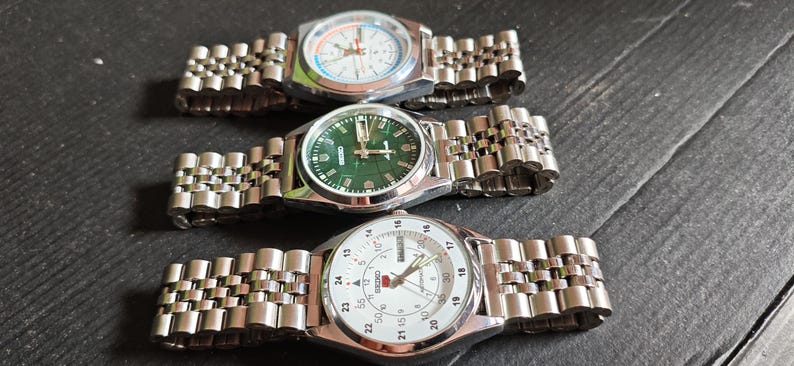 May include: Three stainless steel wristwatches with silver link bands are displayed. The watch faces include a white face with black numbers, a green face with silver hands, and a white face with black numbers and a red logo.