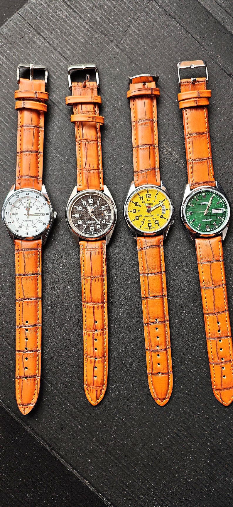 May include: Four wristwatches with orange leather straps and silver watch faces. The watch faces are white, black, yellow, and green. The watch straps have a crocodile skin texture.