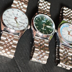 May include: Three stainless steel wristwatches with different colored faces: white, green, and white with blue and orange accents. Each watch has a silver link band. The watches are displayed on a dark surface.