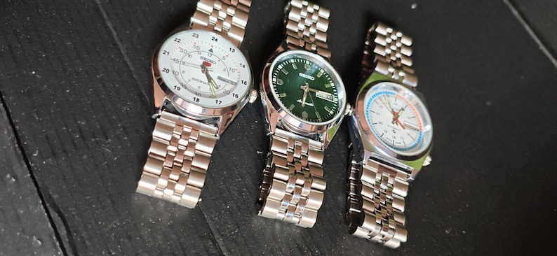 May include: Three stainless steel wristwatches with different colored faces: white, green, and white with blue and orange accents. Each watch has a silver link band. The watches are displayed on a dark surface.