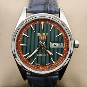 Peut inclure: Une montre automatique Seiko 5 argentée avec un cadran vert et des détails dorés. La montre possède un affichage de la date et un bracelet en cuir bleu. Le cadran présente un motif rayé et un anneau extérieur rouge avec des chiffres.