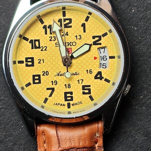 May include: A yellow-faced Seiko automatic wristwatch with a brown leather band. The watch has black numerals and hands, with a date display at the 3 o'clock position. The band is textured.
