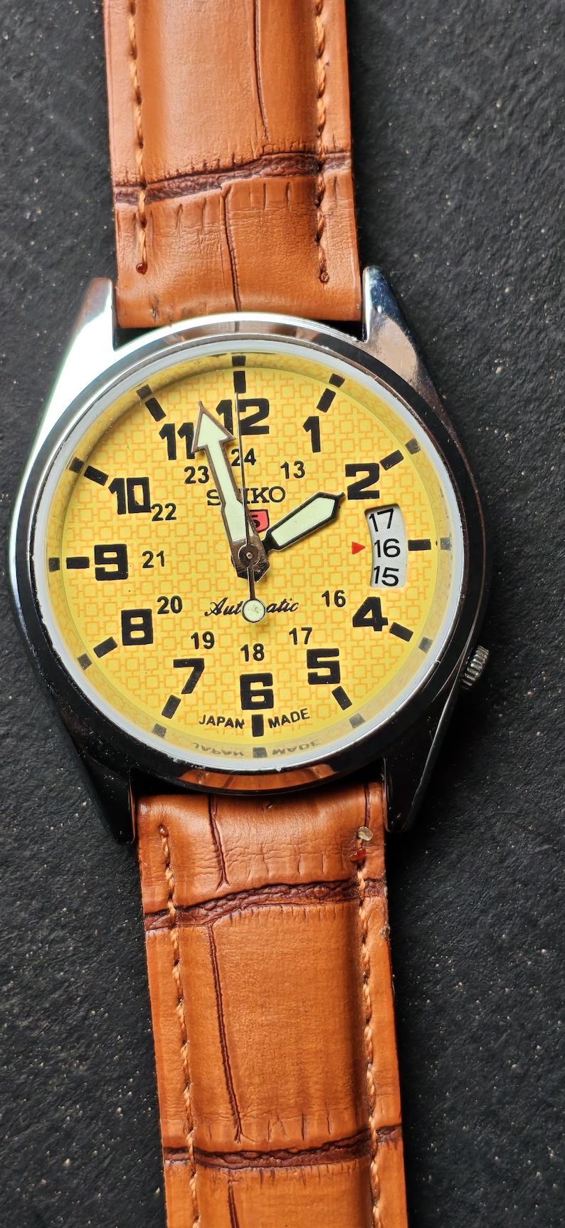 May include: A yellow-faced Seiko automatic wristwatch with a brown leather band. The watch has black numerals and hands, with a date display at the 3 o'clock position. The band is textured.