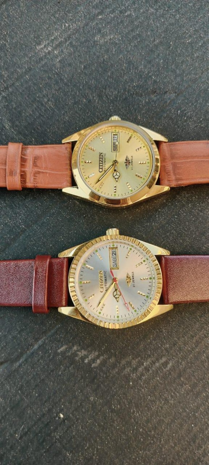 May include: Two Citizen automatic wristwatches with gold-toned cases and brown leather straps. The watch faces are gold and silver, displaying the day and date. The watches have a classic design.