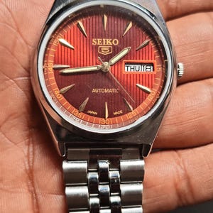 May include: A stainless steel Seiko 5 automatic watch with a red dial. The watch face displays the day and date, with a silver band. The watch is marked "Japan Made".