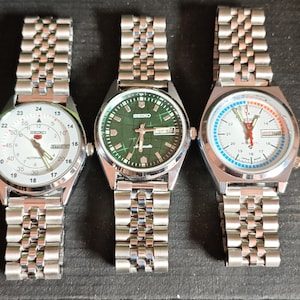 May include: Three Seiko wristwatches with silver-tone metal bands are displayed. The watch faces are white, green, and white with blue accents. Each watch has a different dial design, including date displays and various sub-dials.