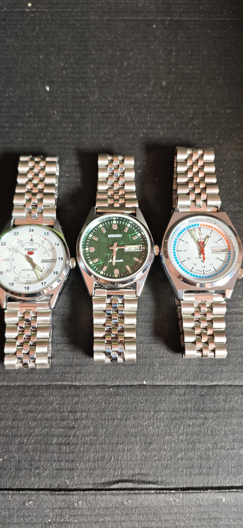May include: Three Seiko wristwatches with silver-tone metal bands are displayed. The watch faces are white, green, and white with blue accents. Each watch has a different dial design, including date displays and various sub-dials.