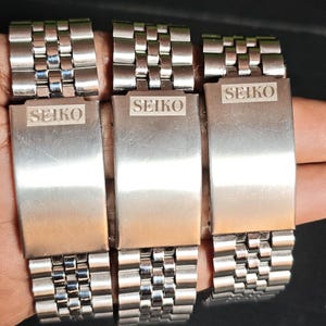 May include: Three stainless steel Seiko watch bands are displayed. Each band has a rectangular clasp with the Seiko logo engraved. The bands are composed of small, interconnected rectangular links, creating a flexible bracelet.