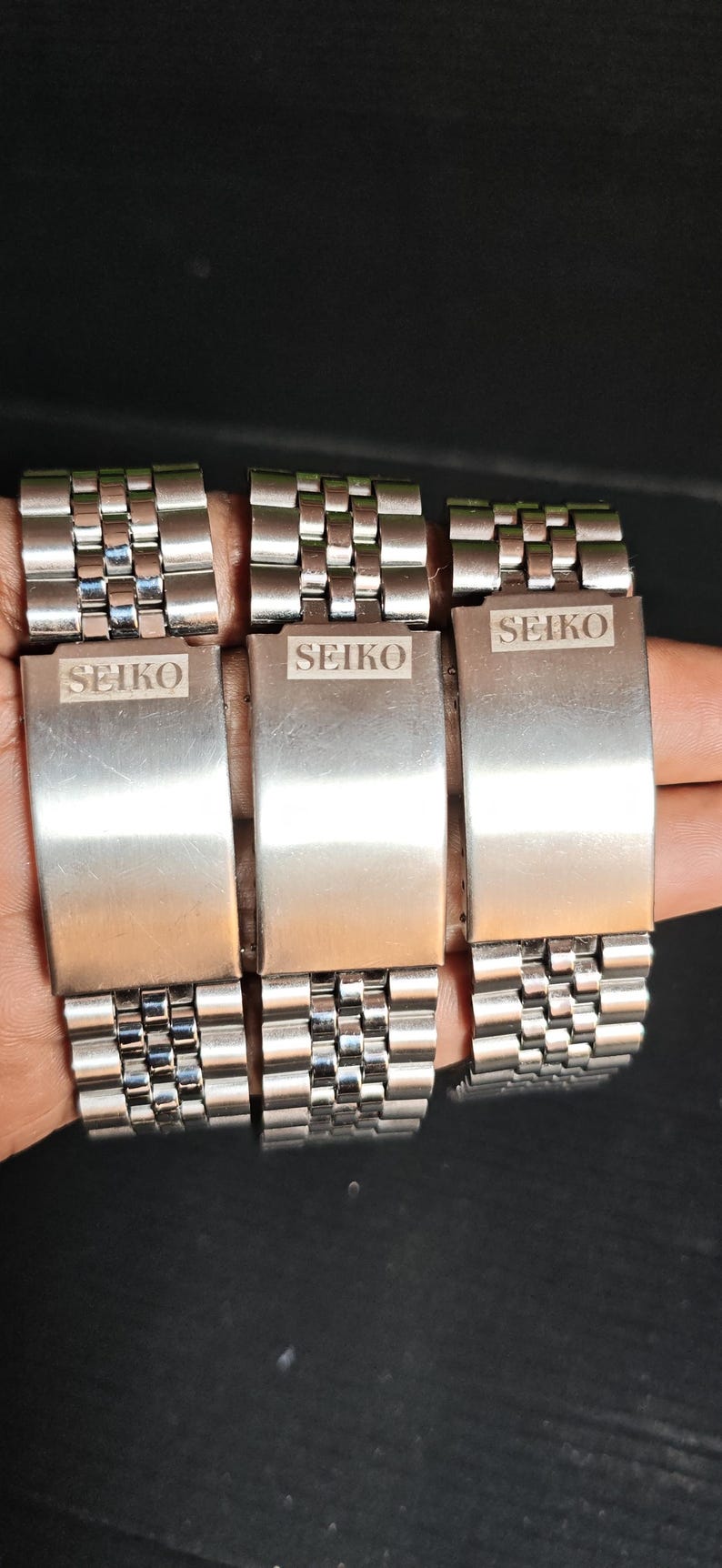 May include: Three stainless steel Seiko watch bands are displayed. Each band has a rectangular clasp with the Seiko logo engraved. The bands are composed of small, interconnected rectangular links, creating a flexible bracelet.