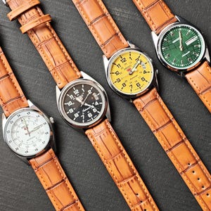 May include: Four wristwatches with brown leather straps are displayed on a dark surface. The watch faces are white, black, yellow, and green. The watches have silver-colored cases and are angled towards the viewer.