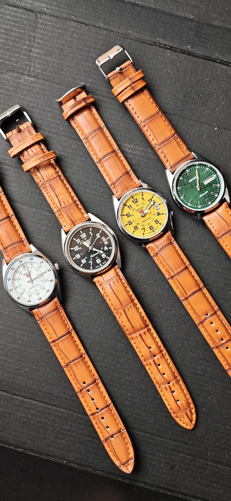 May include: Four wristwatches with brown leather straps are displayed on a dark surface. The watch faces are white, black, yellow, and green. The watches have silver-colored cases and are angled towards the viewer.