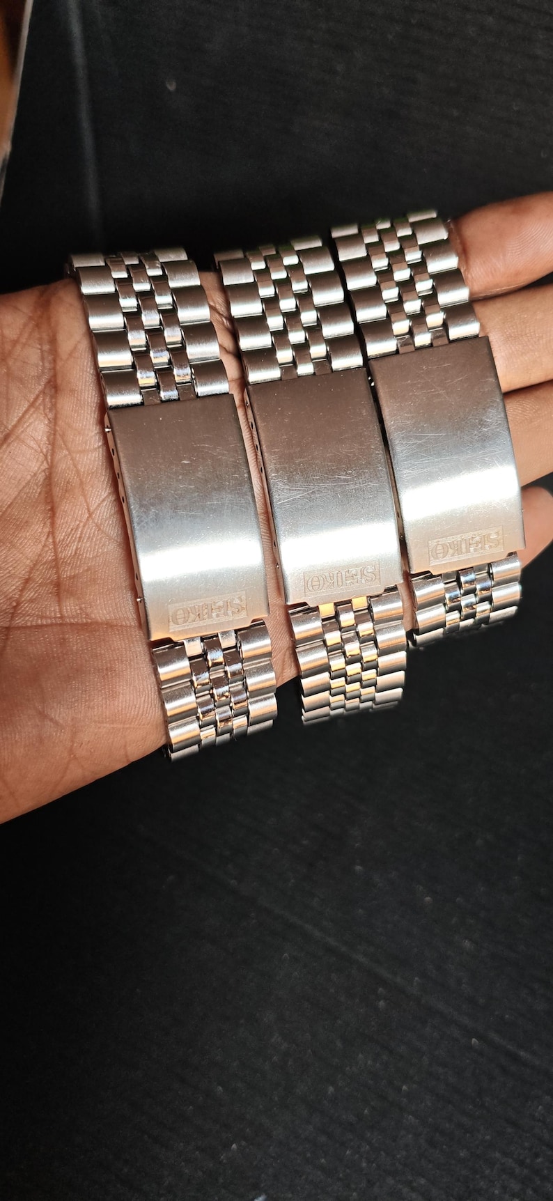 May include: Three stainless steel watch bands with a brushed finish. Each band features a rectangular clasp with the "SEIKO" brand name. The bands are composed of small, interconnected links.