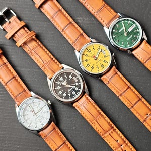 May include: Four wristwatches with brown leather straps and silver-tone cases are displayed. The watch faces are yellow, black, white, and green. The watch straps have a textured, crocodile-like pattern.