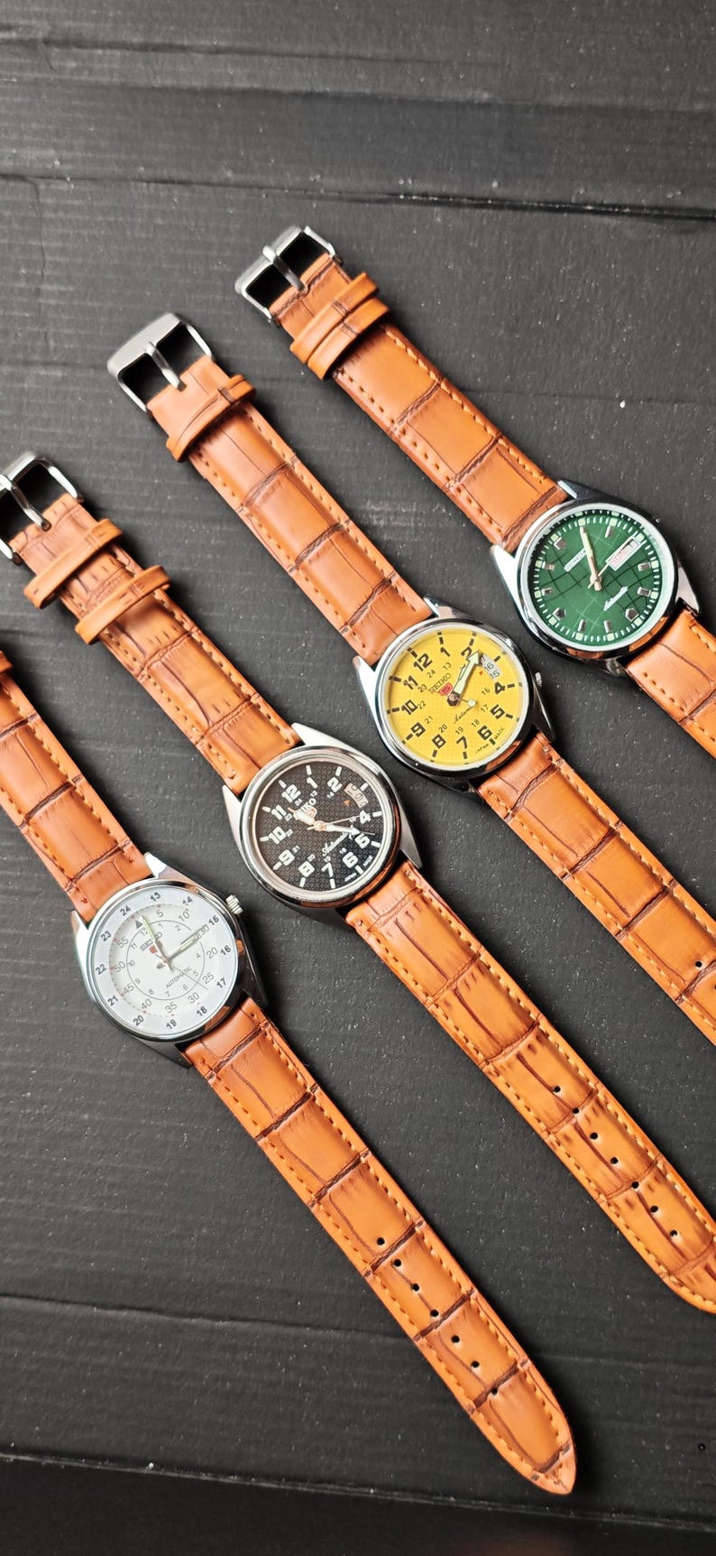 May include: Four wristwatches with brown leather straps and silver-tone cases are displayed. The watch faces are yellow, black, white, and green. The watch straps have a textured, crocodile-like pattern.