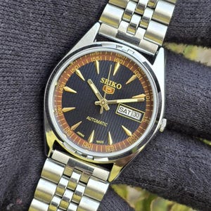 Może przedstawiać: Srebrny automatyczny zegarek na rękę Seiko 5 z czarną tarczą i złotymi wskazówkami i znacznikami. Zegarek posiada wyświetlacz dnia i daty oraz bransoletę ze stali nierdzewnej. Zegarek jest prezentowany na ciemnej powierzchni.