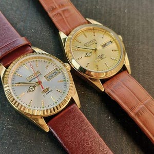 May include: Two gold-toned Citizen automatic watches with brown leather straps. One watch has a silver face with the day and date displayed. The other watch has a gold face. Both watches have gold bezels.