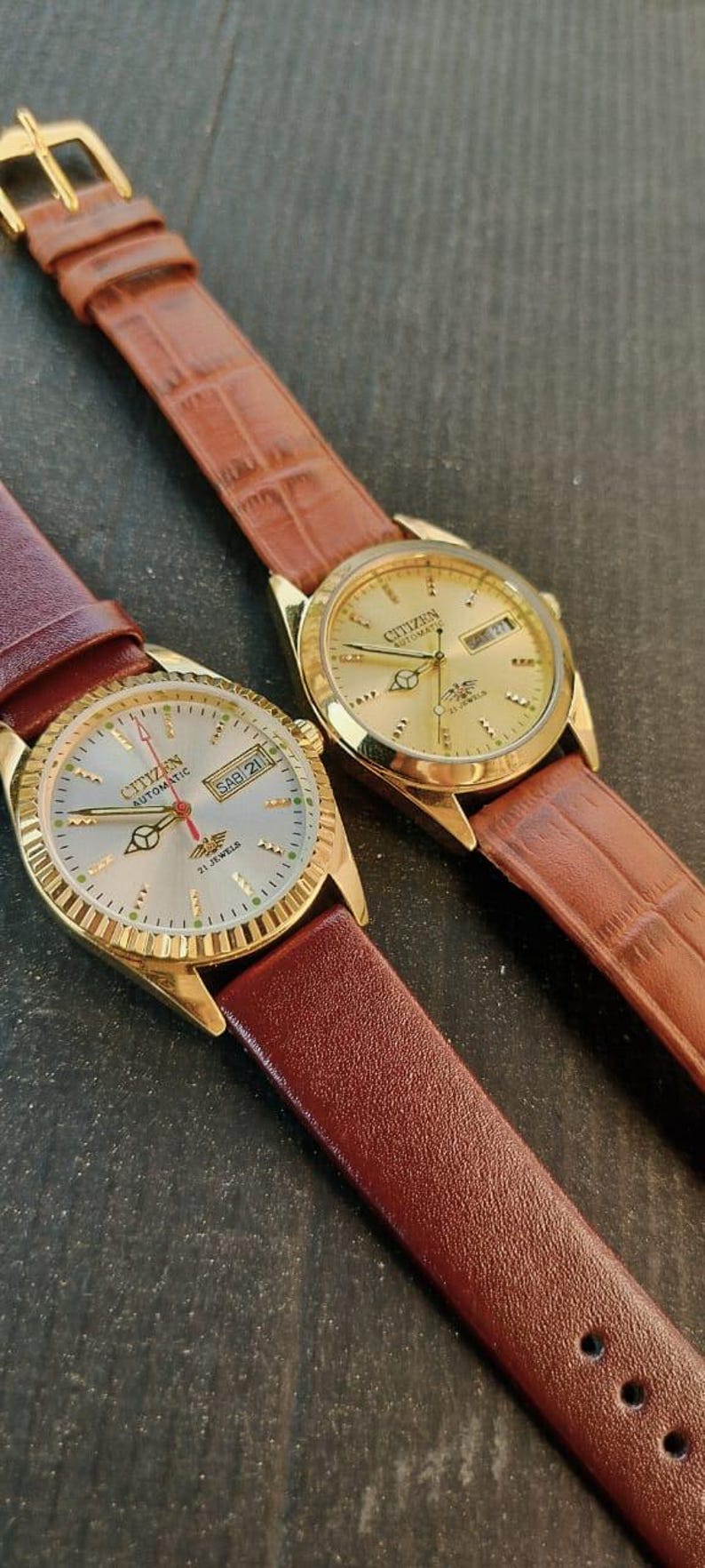 May include: Two gold-toned Citizen automatic watches with brown leather straps. One watch has a silver face with the day and date displayed. The other watch has a gold face. Both watches have gold bezels.
