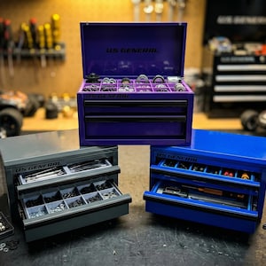 May include: Three small tool chests in purple, gray, and blue, each with drawers open to reveal various tools and parts. The purple chest is open, displaying organized components. The text "US GENERAL" is visible on each chest.