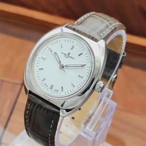 May include: A silver-toned Ulysse Nardin wristwatch with a white face and black leather band. The watch has a round face with silver hands and markers. The band is stitched with a light gray leather on the top and black leather on the bottom.
