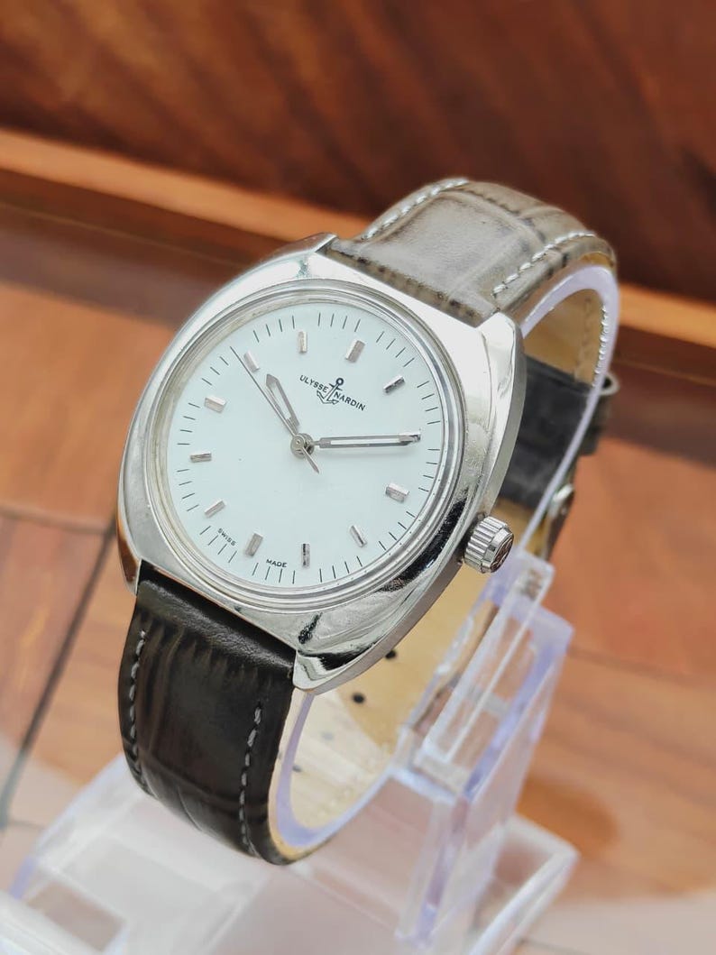 May include: A silver-toned Ulysse Nardin wristwatch with a white face and black leather band. The watch has a round face with silver hands and markers. The band is stitched with a light gray leather on the top and black leather on the bottom.