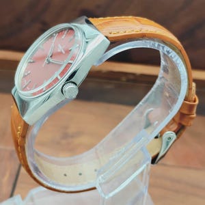 Puede incluir: Un reloj de pulsera vintage con caja plateada y esfera color salm&oacute;n. El reloj presenta una correa de cuero marr&oacute;n. El reloj se exhibe sobre un soporte de pl&aacute;stico transparente, mostrando su dise&ntilde;o cl&aacute;sico y artesan&iacute;a.