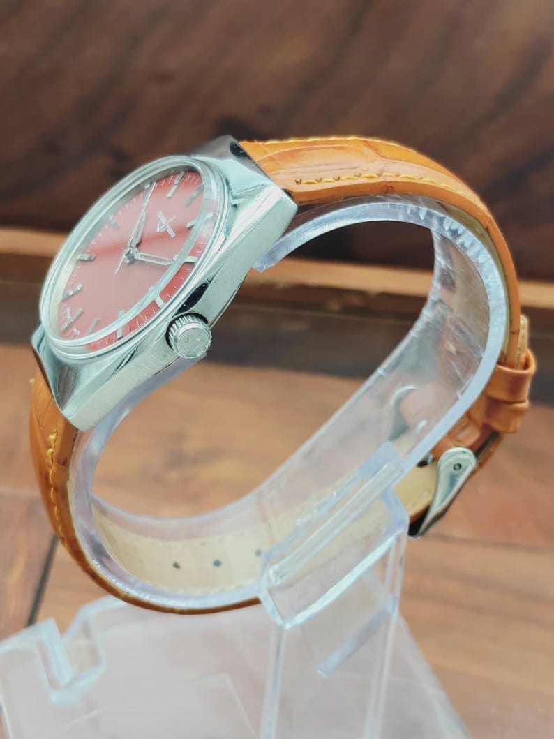 Puede incluir: Un reloj de pulsera vintage con caja plateada y esfera color salm&oacute;n. El reloj presenta una correa de cuero marr&oacute;n. El reloj se exhibe sobre un soporte de pl&aacute;stico transparente, mostrando su dise&ntilde;o cl&aacute;sico y artesan&iacute;a.