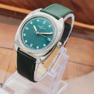K&ouml;nnte beinhalten: Eine silberne Armbanduhr mit gr&uuml;nem Zifferblatt und gr&uuml;nem Lederarmband. Das Zifferblatt zeigt den Markennamen "ULYSSE NARDIN" und wei&szlig;e Stundenmarkierungen. Das Armband ist mit gr&uuml;nem Faden gen&auml;ht.