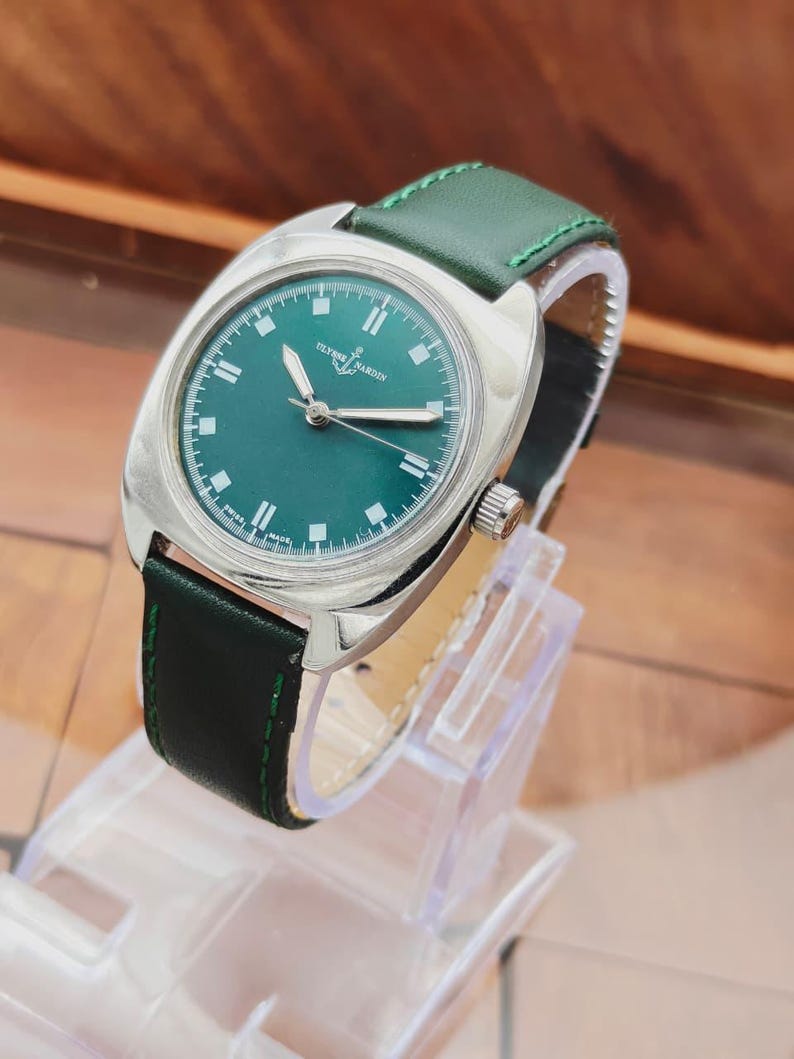 K&ouml;nnte beinhalten: Eine silberne Armbanduhr mit gr&uuml;nem Zifferblatt und gr&uuml;nem Lederarmband. Das Zifferblatt zeigt den Markennamen "ULYSSE NARDIN" und wei&szlig;e Stundenmarkierungen. Das Armband ist mit gr&uuml;nem Faden gen&auml;ht.
