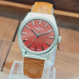 Puede incluir: Un reloj de pulsera plateado con esfera roja y correa de cuero marr&oacute;n. La esfera del reloj presenta marcadores de hora plateados y la marca "Ulysse Nardin". La correa est&aacute; cosida con hilo marr&oacute;n claro.