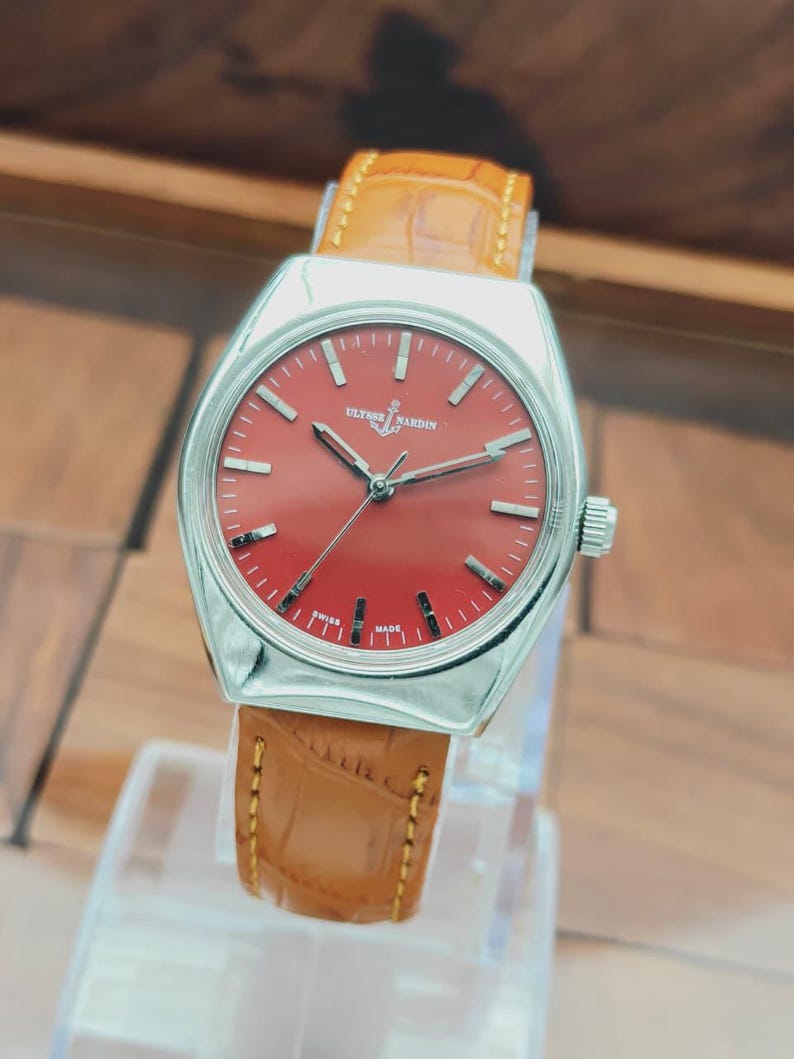 Puede incluir: Un reloj de pulsera plateado con esfera roja y correa de cuero marr&oacute;n. La esfera del reloj presenta marcadores de hora plateados y la marca "Ulysse Nardin". La correa est&aacute; cosida con hilo marr&oacute;n claro.