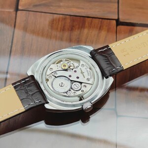Puede incluir: Un reloj de pulsera plateado con una correa de cuero marr&oacute;n. La parte trasera del reloj est&aacute; abierta, revelando los intrincados mecanismos internos, incluyendo engranajes y joyas. La correa est&aacute; sujeta con cuero marr&oacute;n oscuro y tiene un reverso beige claro.