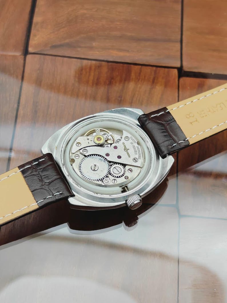 Puede incluir: Un reloj de pulsera plateado con una correa de cuero marr&oacute;n. La parte trasera del reloj est&aacute; abierta, revelando los intrincados mecanismos internos, incluyendo engranajes y joyas. La correa est&aacute; sujeta con cuero marr&oacute;n oscuro y tiene un reverso beige claro.