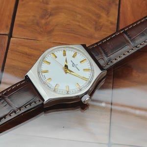 May include: A silver wristwatch with a white face and gold hands and markers. The watch has a brown leather strap. The watch face displays the brand name "Ulysse Nardin".