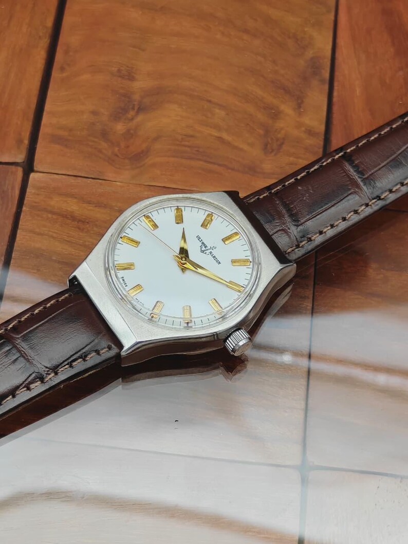 May include: A silver wristwatch with a white face and gold hands and markers. The watch has a brown leather strap. The watch face displays the brand name "Ulysse Nardin".