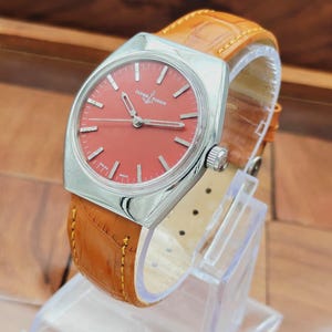 Puede incluir: Un reloj Ulysse Nardin vintage con esfera roja y caja plateada. El reloj tiene una correa de cuero marr&oacute;n con un patr&oacute;n de cocodrilo. El reloj se exhibe en un soporte de pl&aacute;stico transparente.