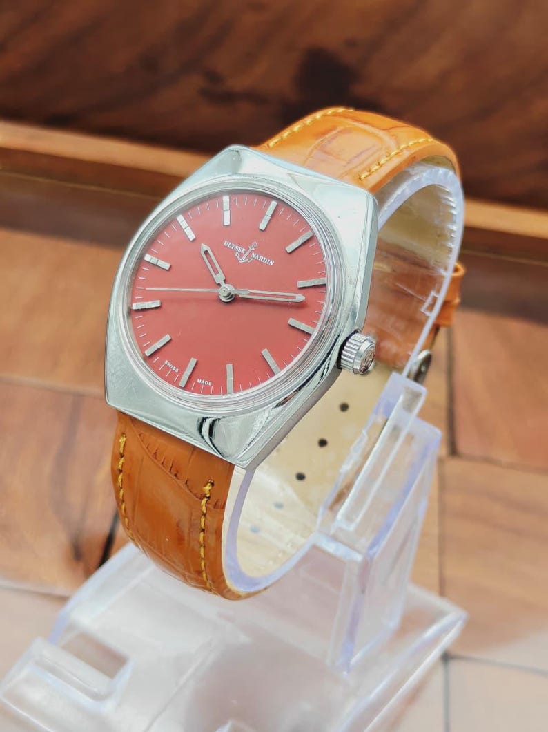 Puede incluir: Un reloj Ulysse Nardin vintage con esfera roja y caja plateada. El reloj tiene una correa de cuero marr&oacute;n con un patr&oacute;n de cocodrilo. El reloj se exhibe en un soporte de pl&aacute;stico transparente.