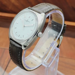 May include: A silver-toned wristwatch with a white face and a brown leather band. The watch has a round face with silver hands and markers. The band is dark brown with a textured pattern. The watch is displayed on a clear plastic stand.