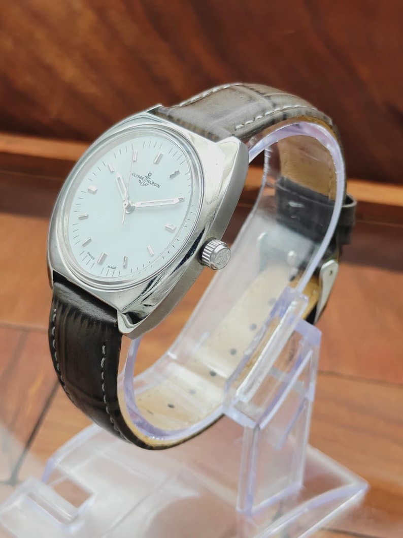 May include: A silver-toned wristwatch with a white face and a brown leather band. The watch has a round face with silver hands and markers. The band is dark brown with a textured pattern. The watch is displayed on a clear plastic stand.