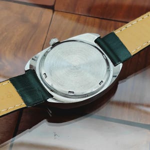 K&ouml;nnte beinhalten: Nahaufnahme der R&uuml;ckseite einer silberfarbenen Armbanduhr mit strukturiertem, kreisf&ouml;rmigem Design. Die Uhr hat ein schwarzes Lederarmband mit gelber Unterseite. Die Krone der Uhr ist sichtbar, auf einer Holzoberfl&auml;che.