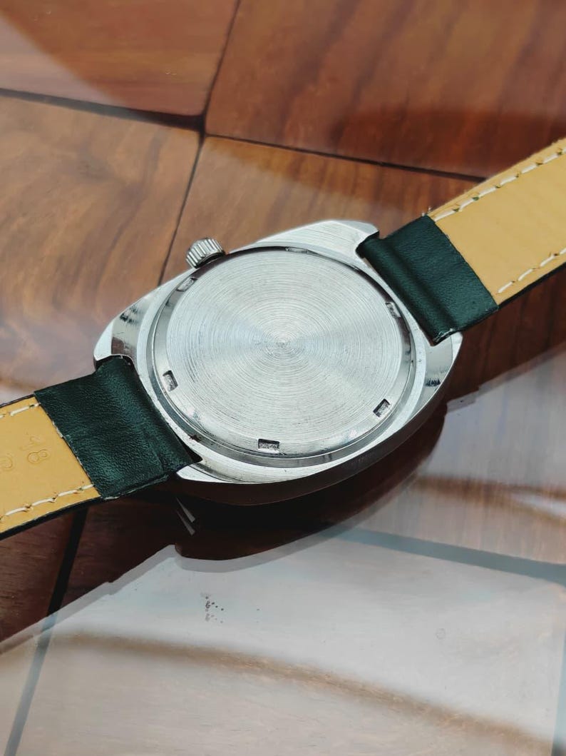 K&ouml;nnte beinhalten: Nahaufnahme der R&uuml;ckseite einer silberfarbenen Armbanduhr mit strukturiertem, kreisf&ouml;rmigem Design. Die Uhr hat ein schwarzes Lederarmband mit gelber Unterseite. Die Krone der Uhr ist sichtbar, auf einer Holzoberfl&auml;che.
