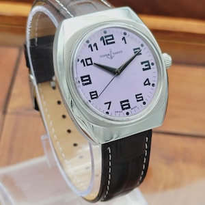 Puede incluir: Un reloj de pulsera plateado con esfera rosa claro, n&uacute;meros negros y manecillas negras. El reloj tiene una correa de cuero negro con costuras blancas. La marca "ULYSSE NARDIN" es visible en la esfera. El reloj se muestra sobre un soporte transparente.