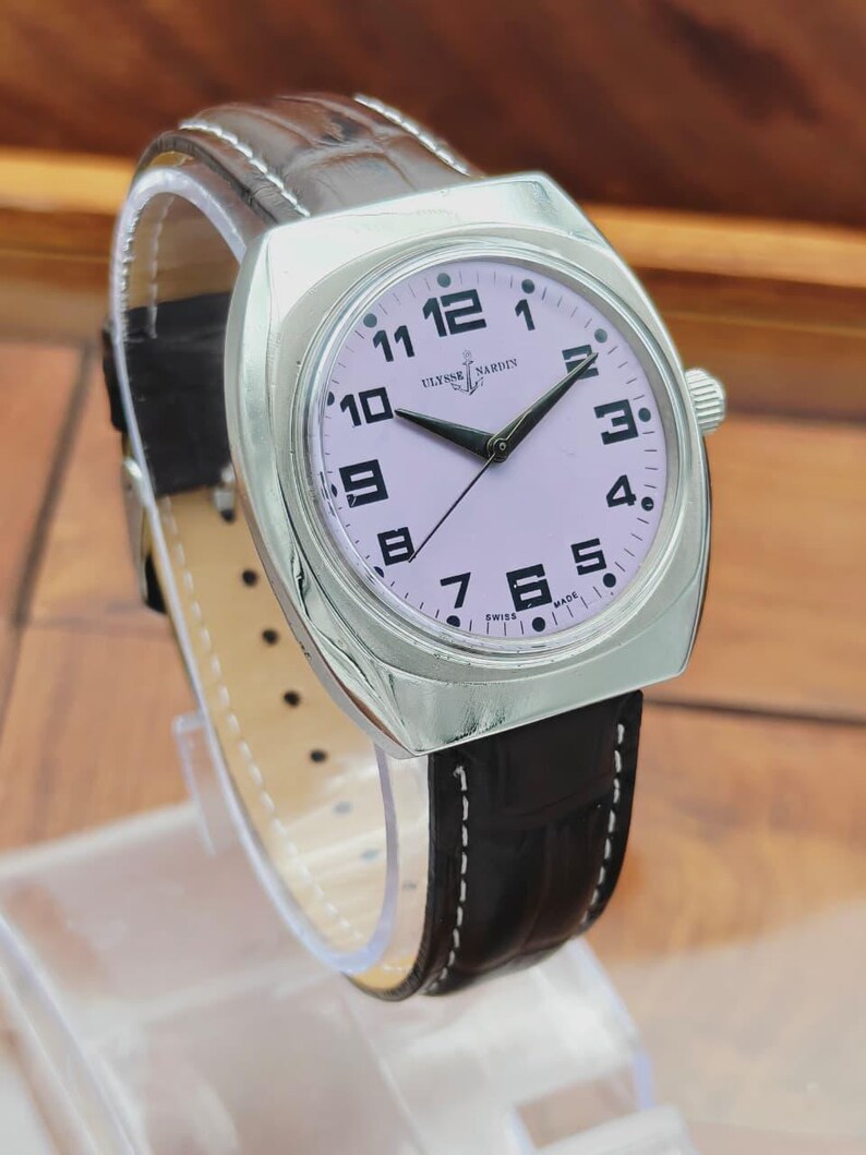 Puede incluir: Un reloj de pulsera plateado con esfera rosa claro, n&uacute;meros negros y manecillas negras. El reloj tiene una correa de cuero negro con costuras blancas. La marca "ULYSSE NARDIN" es visible en la esfera. El reloj se muestra sobre un soporte transparente.